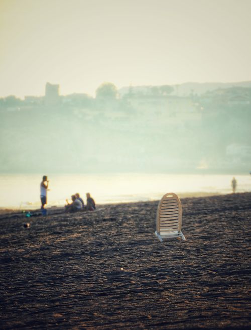 Free Tranquil beach scene featuring an empty chair and people enjoying the serene sunset by the ocean. Stock Photo