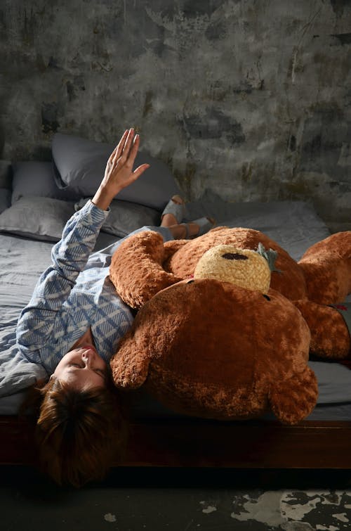 Free Woman in Gray Long Sleeve Shirt Lying on Bed Beside Brown Bear Plush Toy Stock Photo