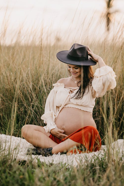 Free Smiling pregnant woman in hat looking down while sitting on grass in countryside Stock Photo