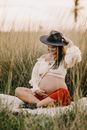 Smiling pregnant woman in hat looking down while sitting on grass in countryside