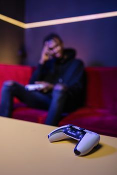 Focus on a video game controller with a blurred player in the background on a red sofa.