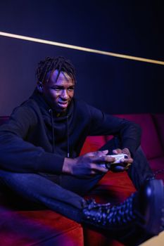 Young man intensely playing video games with controller on a red couch in a dimly lit room.