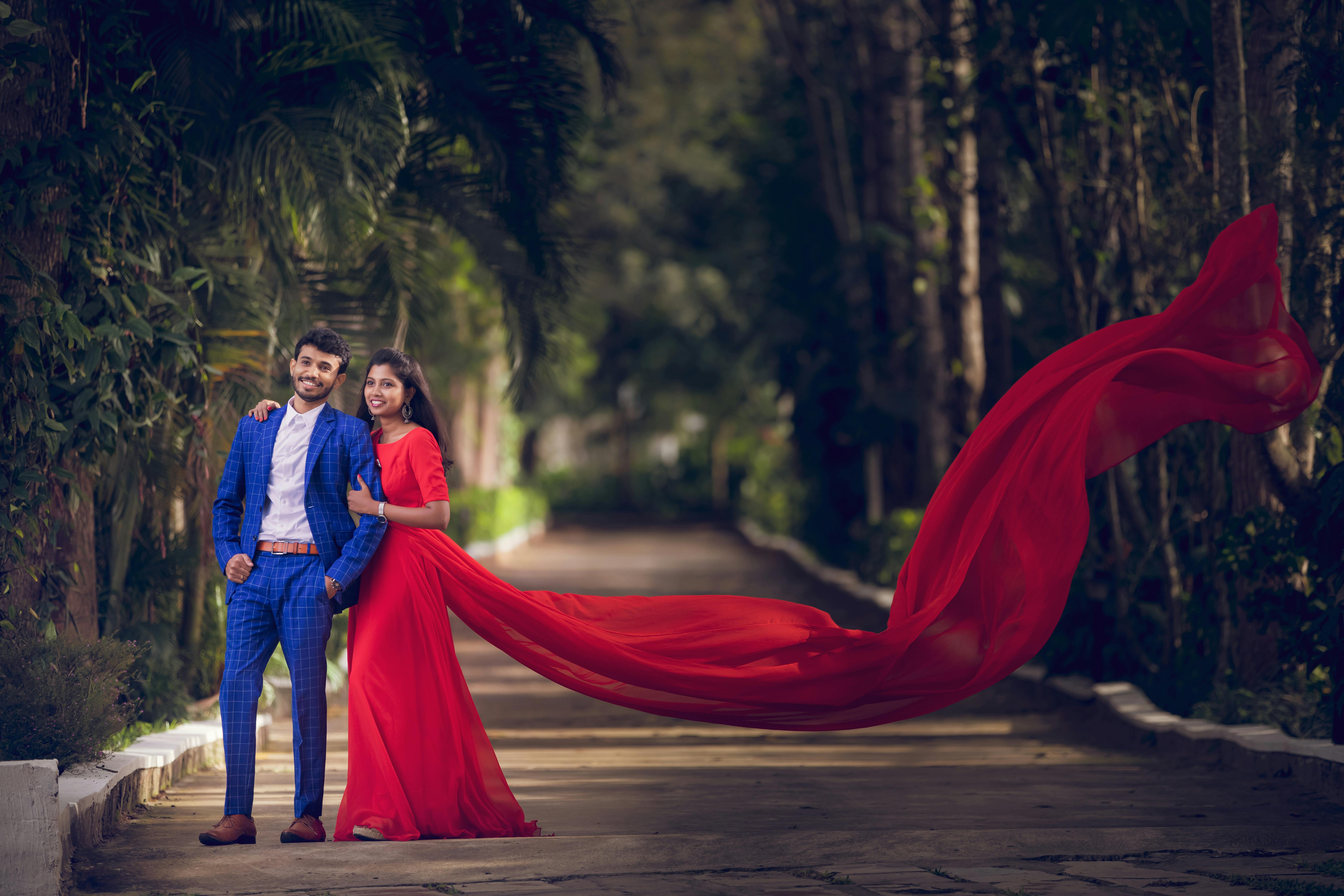 Happy couple in elegant attire posing outdoors with flowing red fabric and lush garden background.