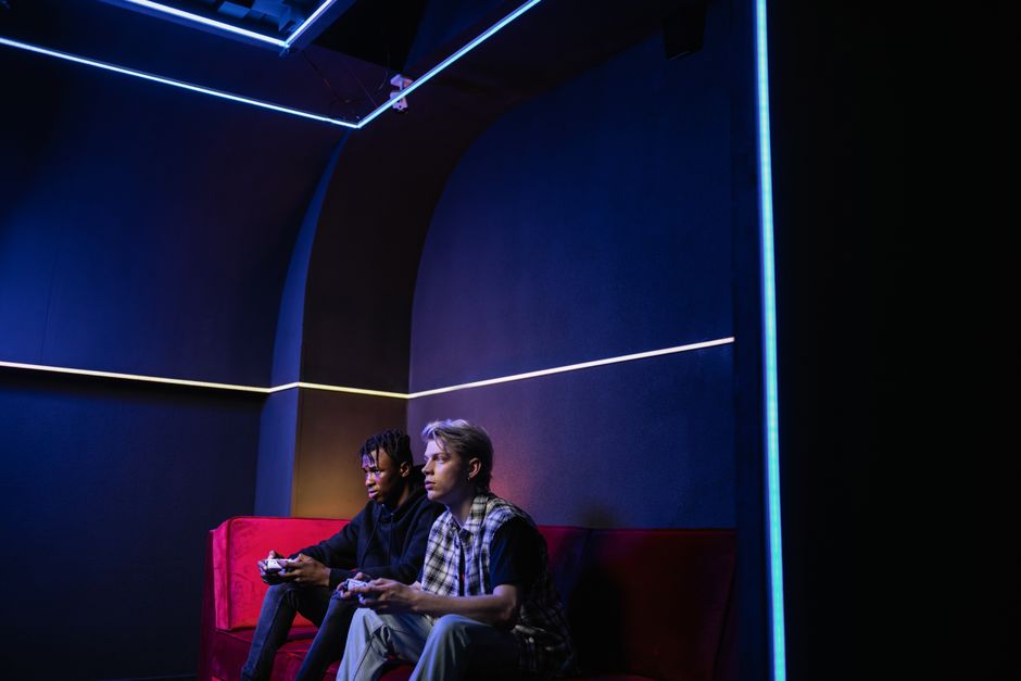 Two men engaged in video gaming on a couch with neon lighting in a dark room.