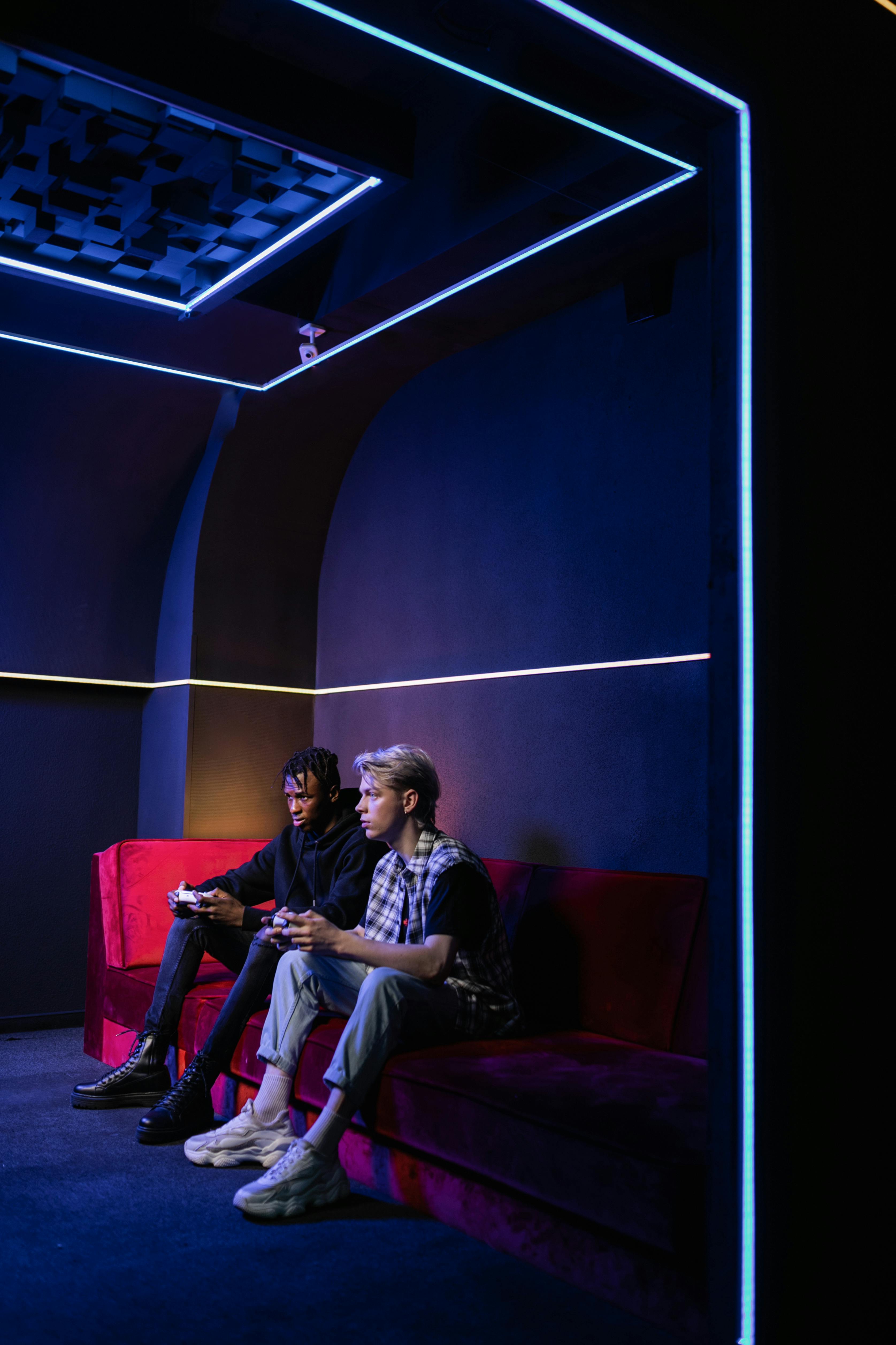 Two young men with controllers gaming in a neon-lit room on a red couch.