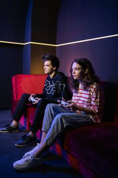 A man and a woman focused on playing video games together on a red couch in a dimly lit room.