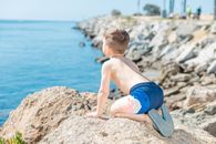 Topless Boy in Blue Shorts Sitting on Rock Near Sea