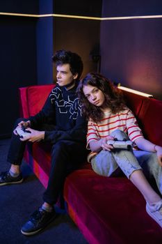 Teenagers sitting on a red sofa intently playing video games indoors.