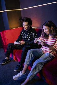 Two teenagers playing video games on a vibrant red sofa indoors.