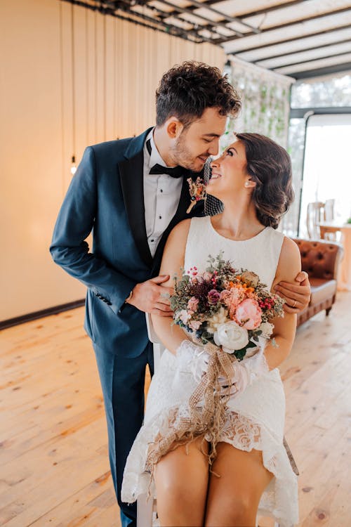 Happy young newlywed bride holding bouquet of flowers wearing elegant wedding dress sitting next to groom in suit smiling and looking at each other