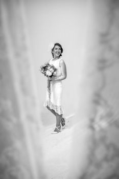 Black and white full body of cheerful young newlywed female wearing elegant white wedding dress and holding flowers in hands while looking away