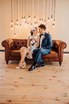 Bride and groom lovingly glance at each other on a leather sofa under pendant lights. Elegant wedding setting.