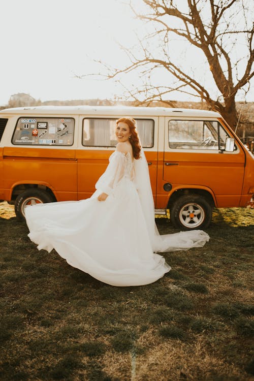 Free Full body young happy female in long white wedding dress standing on grassy meadow in front of red old fashioned camper and looking in camera over shoulder in sunlight Stock Photo