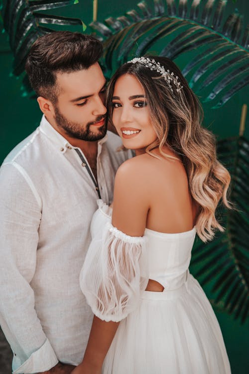 Delighted smiling young bride in white dress holding hands with handsome groom in white shirt while standing together near palm leaves and looking at camera