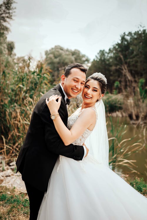 Happy laughing newlywed couple wearing wedding clothes standing near lake in nature while embracing and looking at camera