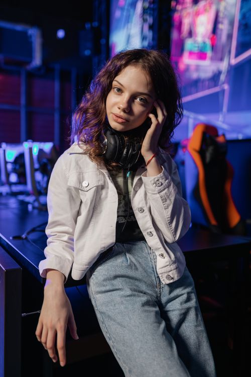Free Portrait of a young woman in a gaming environment, wearing a headset and casual attire, with colorful ambient lights. Stock Photo
