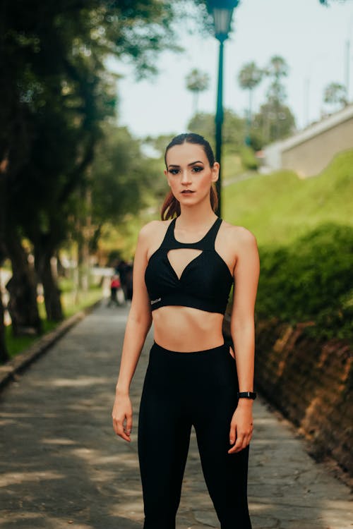 Confident fit female wearing black leggings and top standing on park alley near green lawn on sunny summer day