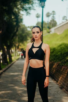 Confident fit female wearing black leggings and top standing on park alley near green lawn on sunny summer day