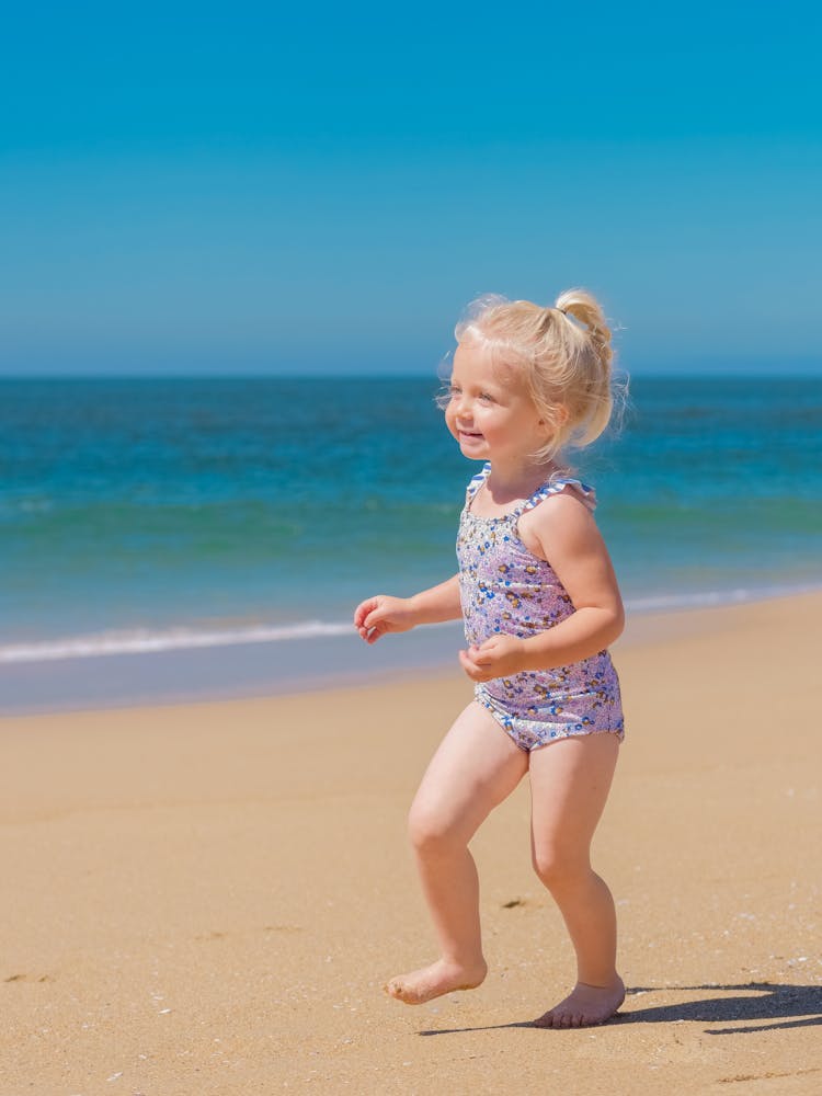 A Cute Young Girl In Floral Swimsuit Walking On The Beach Sand
