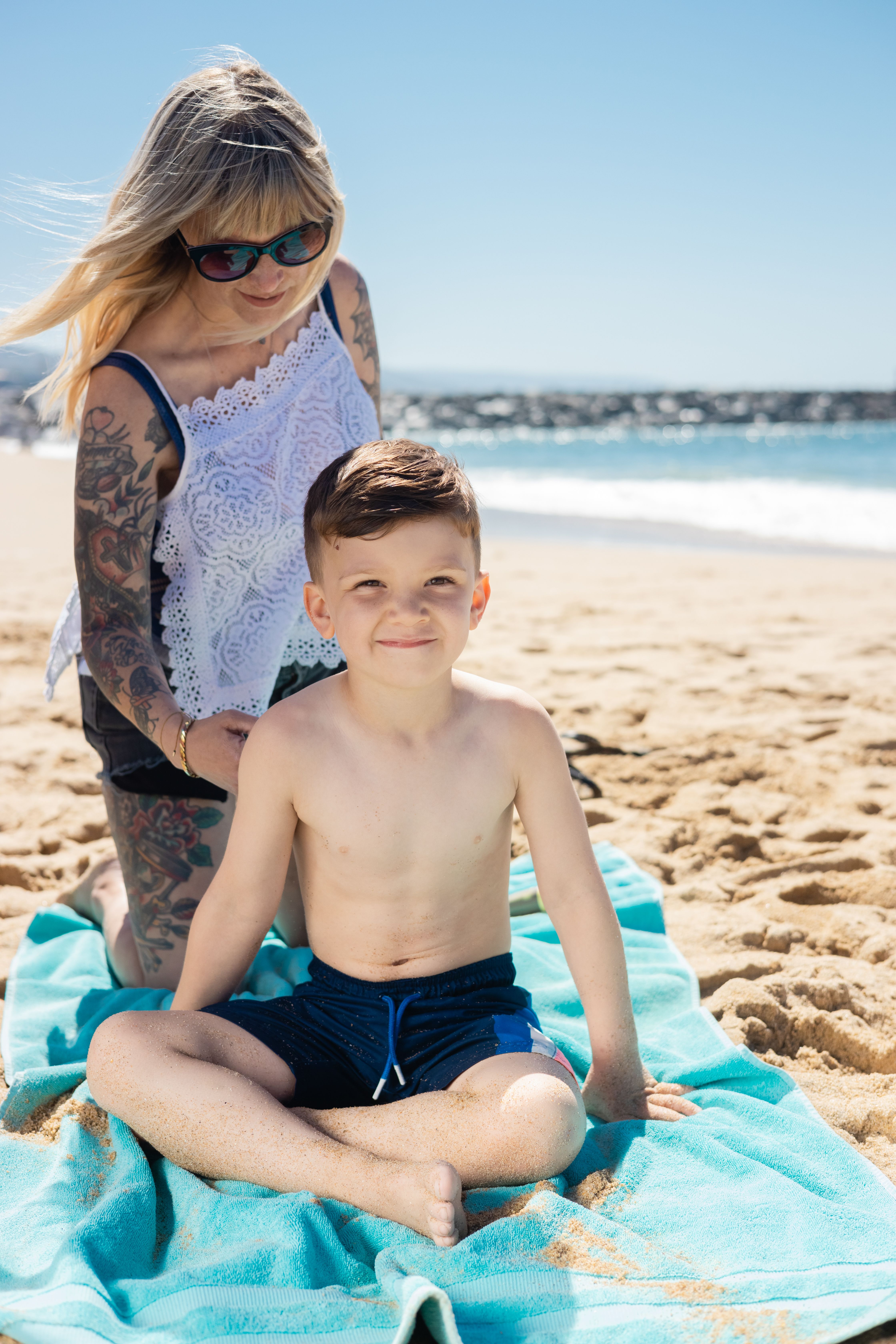 A mother and her son enjoy quality time together on a sunny beach with clear skies and gentle waves.