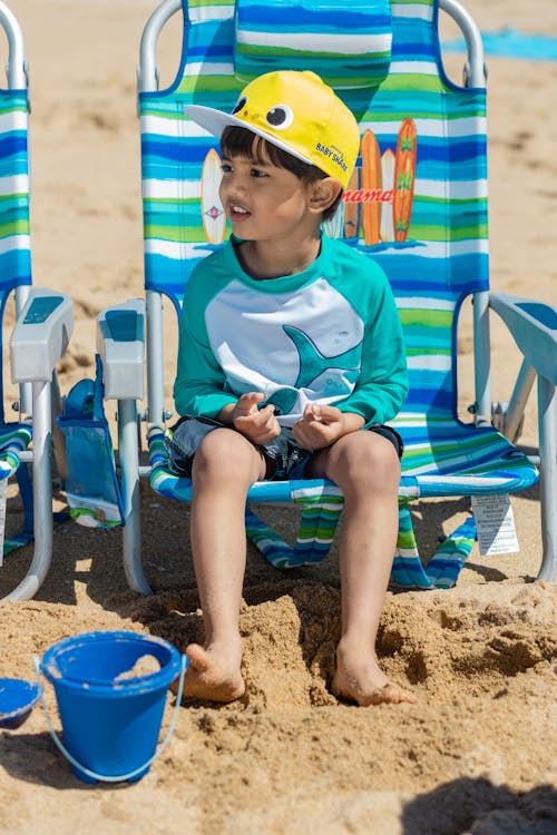 A Young Boy Sitting on a Beach Chair while Wearing a Yellow Cap