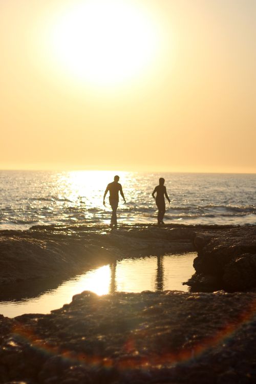 Free Two people walking on a rocky beach silhouette against a golden sunset over the sea. Stock Photo