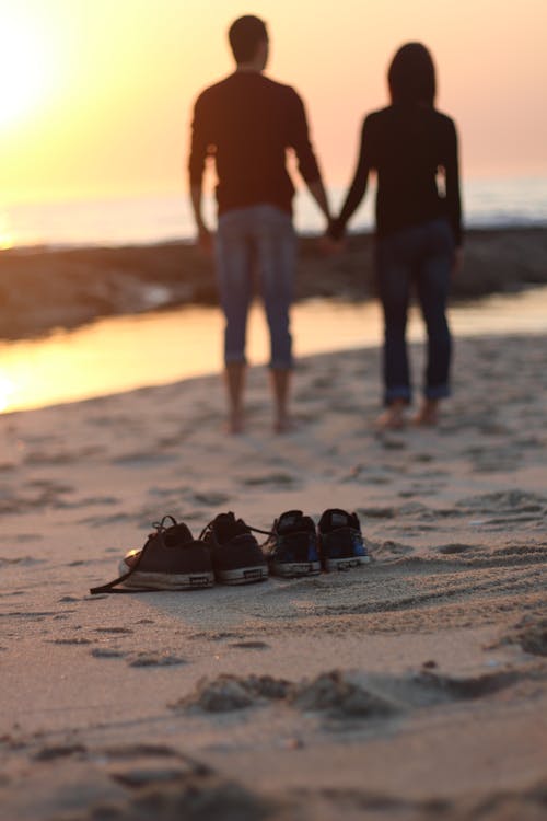 Two Pair of Sneakers Near Couple Standing on Beach Sand