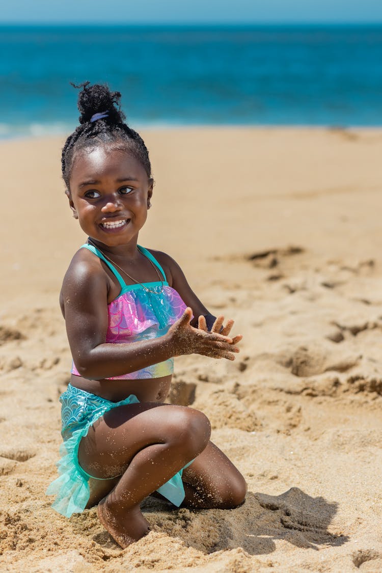 Little Girl In Pink And Blue Bikini Squatting On Beach Sand