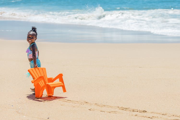 Cute Girl In Swimsuit Pulling A Chair On Beach Sand