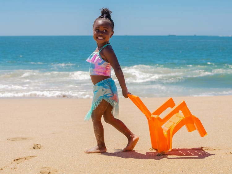 A Young Girl In Swimsuit Pulling A Chair On The Beach