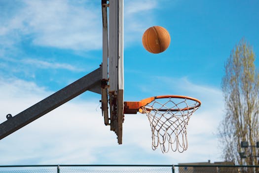Basketball mid-air nearing the hoop on an outdoor court under a clear blue sky.