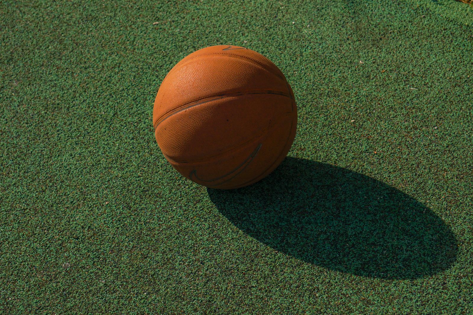 Close de bola de basquete apoiada no piso da quadra, com sombra marcada pelo sol