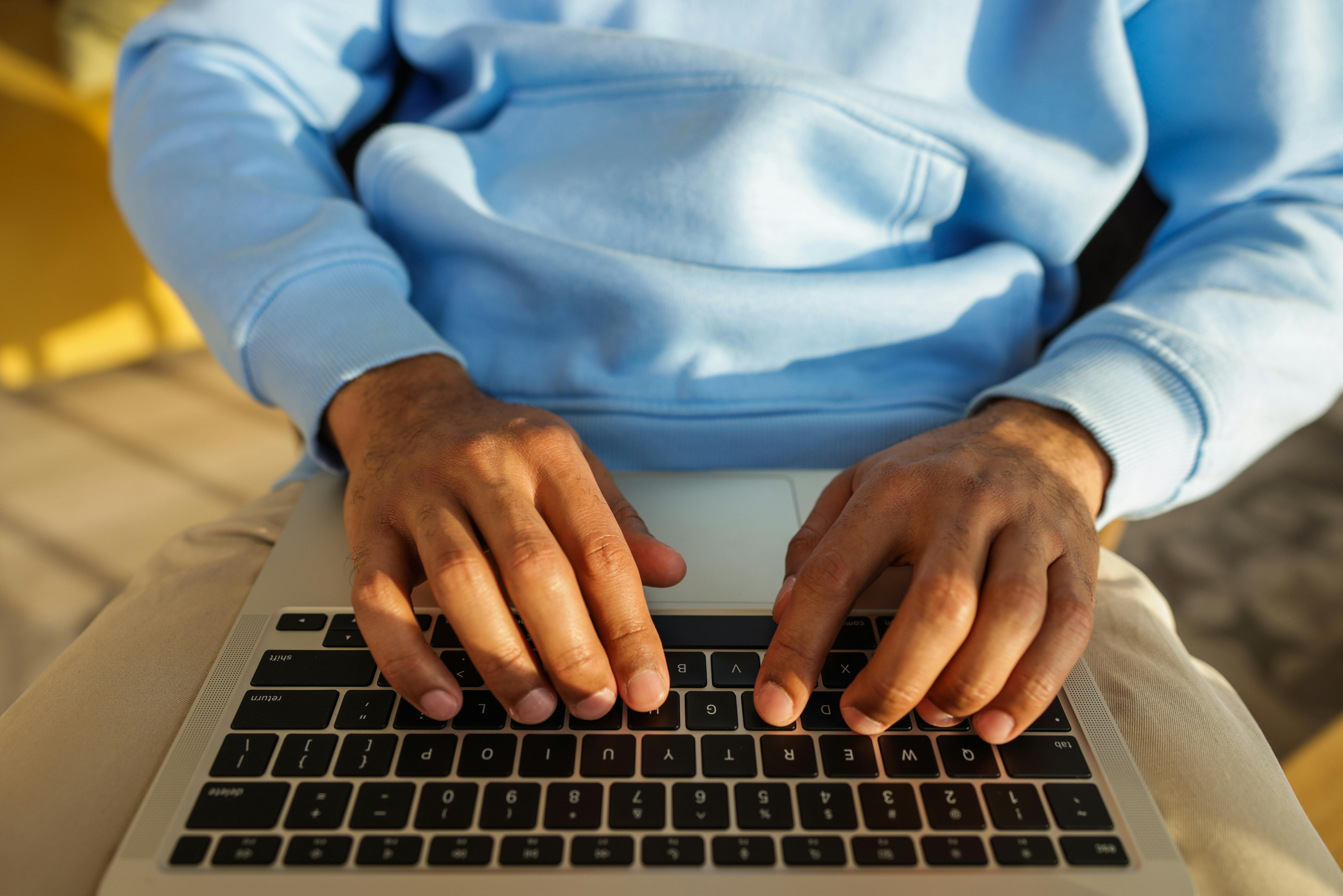 Hands in a blue hoodie typing on a laptop keyboard, indoors, in bright light.