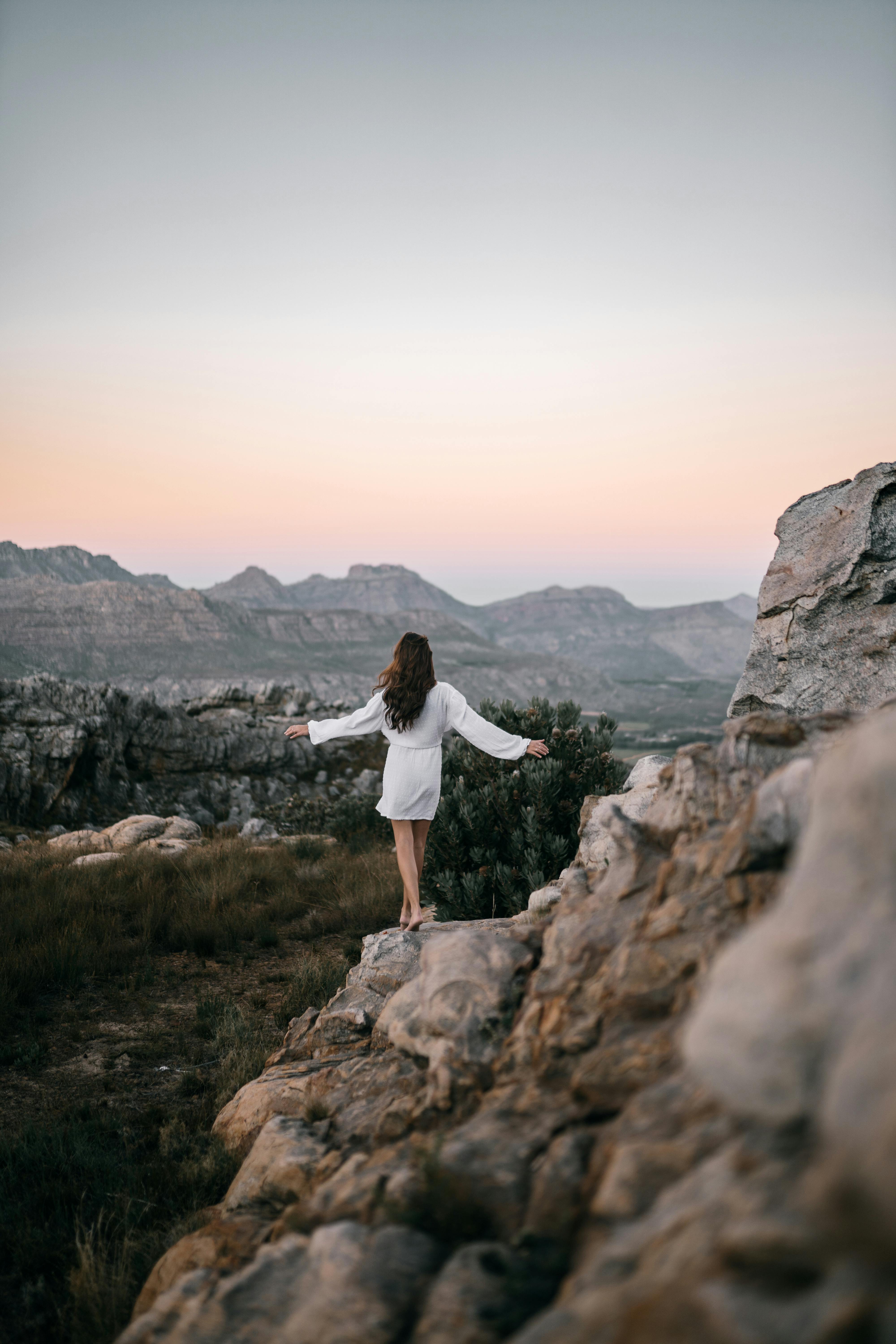 身穿白色连衣裙的女子站在岩石地形上，在日落时拥抱大自然