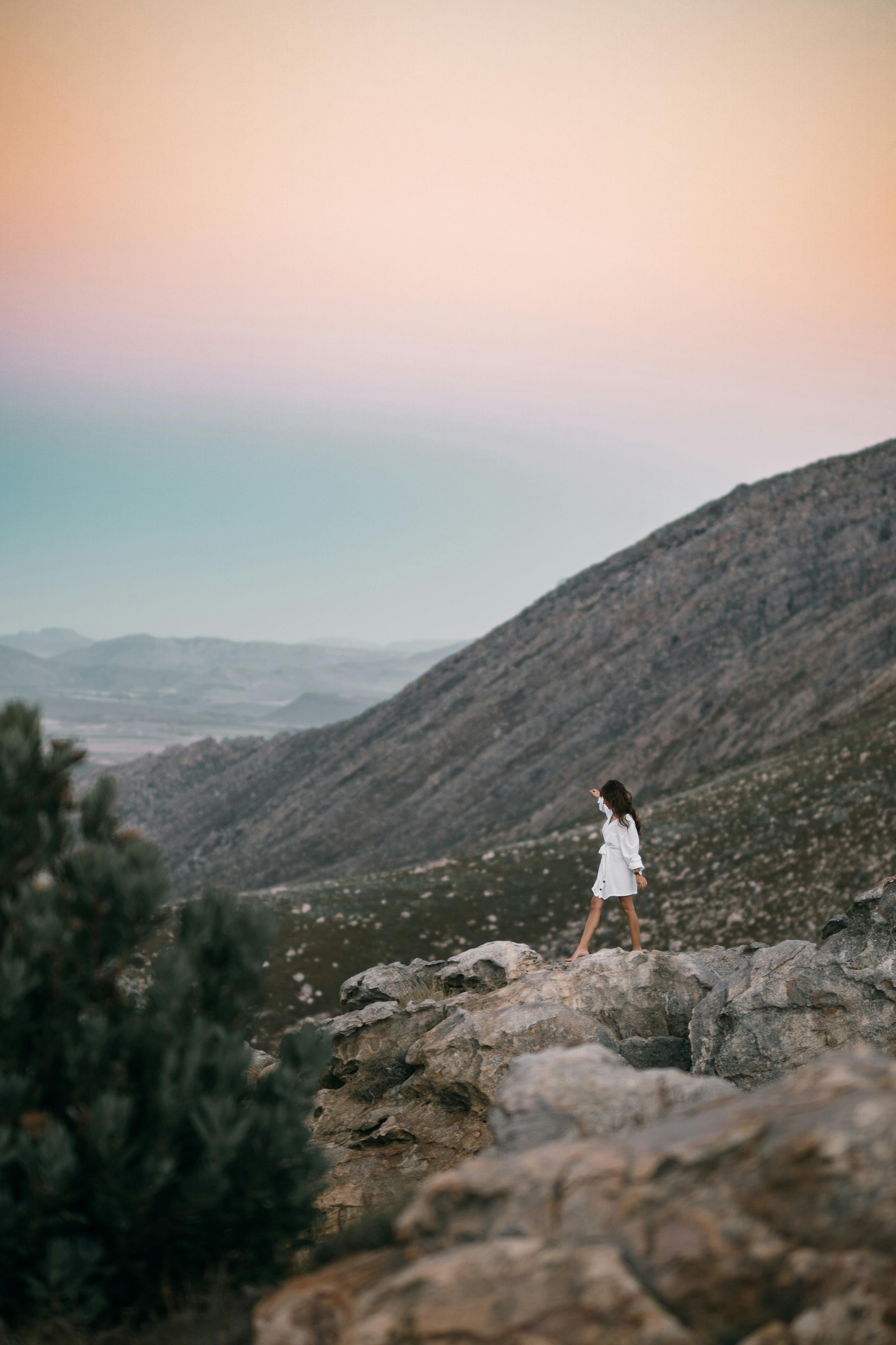 A woman in white walks on rocky terrain during a serene sunset with mountains in the background.