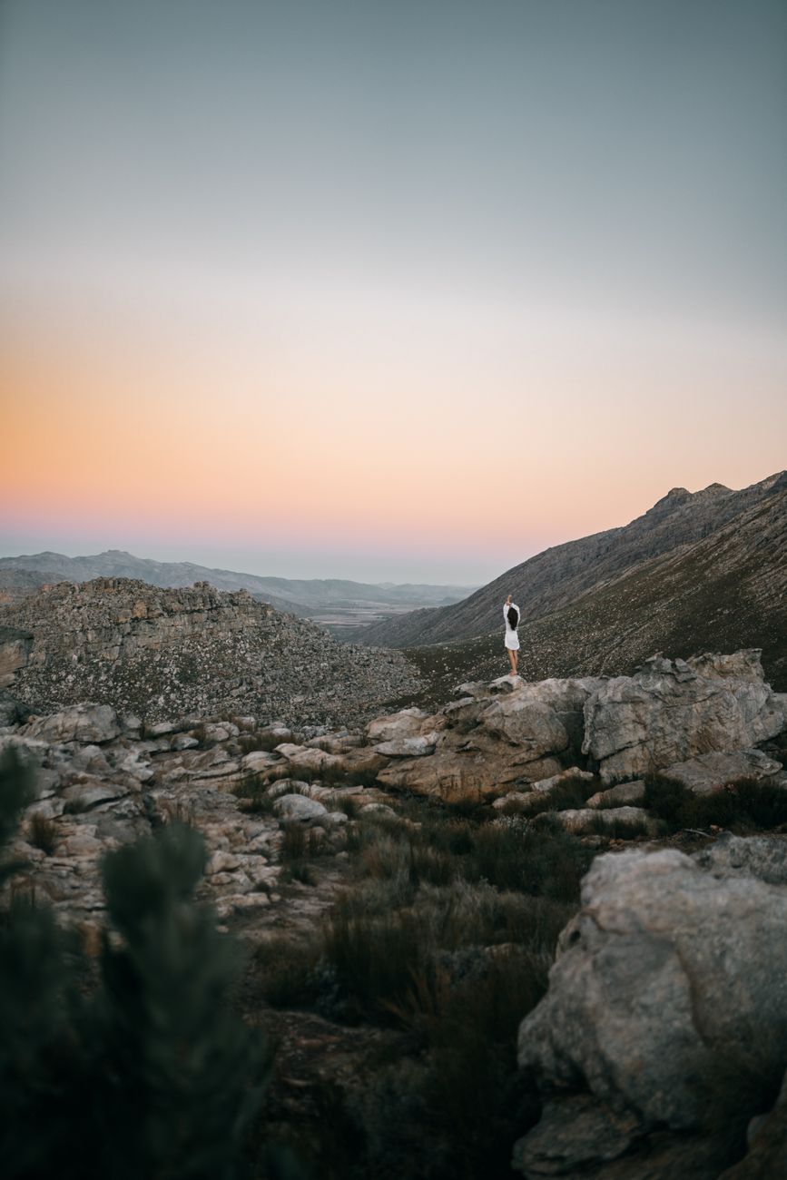 A lone woman in a white dress stands on a rocky mountain during sunset, capturing the sense of solitude and wanderlust.