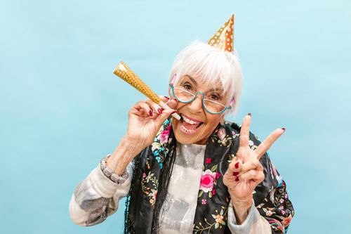 Free Senior woman in party hat joyfully celebrating, making peace sign on a light blue background. Stock Photo