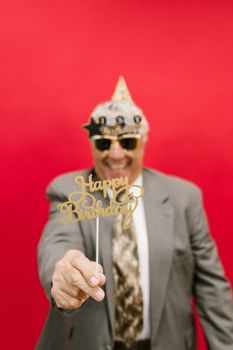 Smiling man in festive attire holding a 'Happy Birthday' sign against a red backdrop.