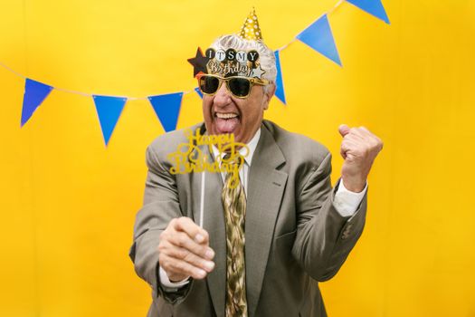 Elderly man joyfully celebrates his birthday with playful props and a bright yellow backdrop.