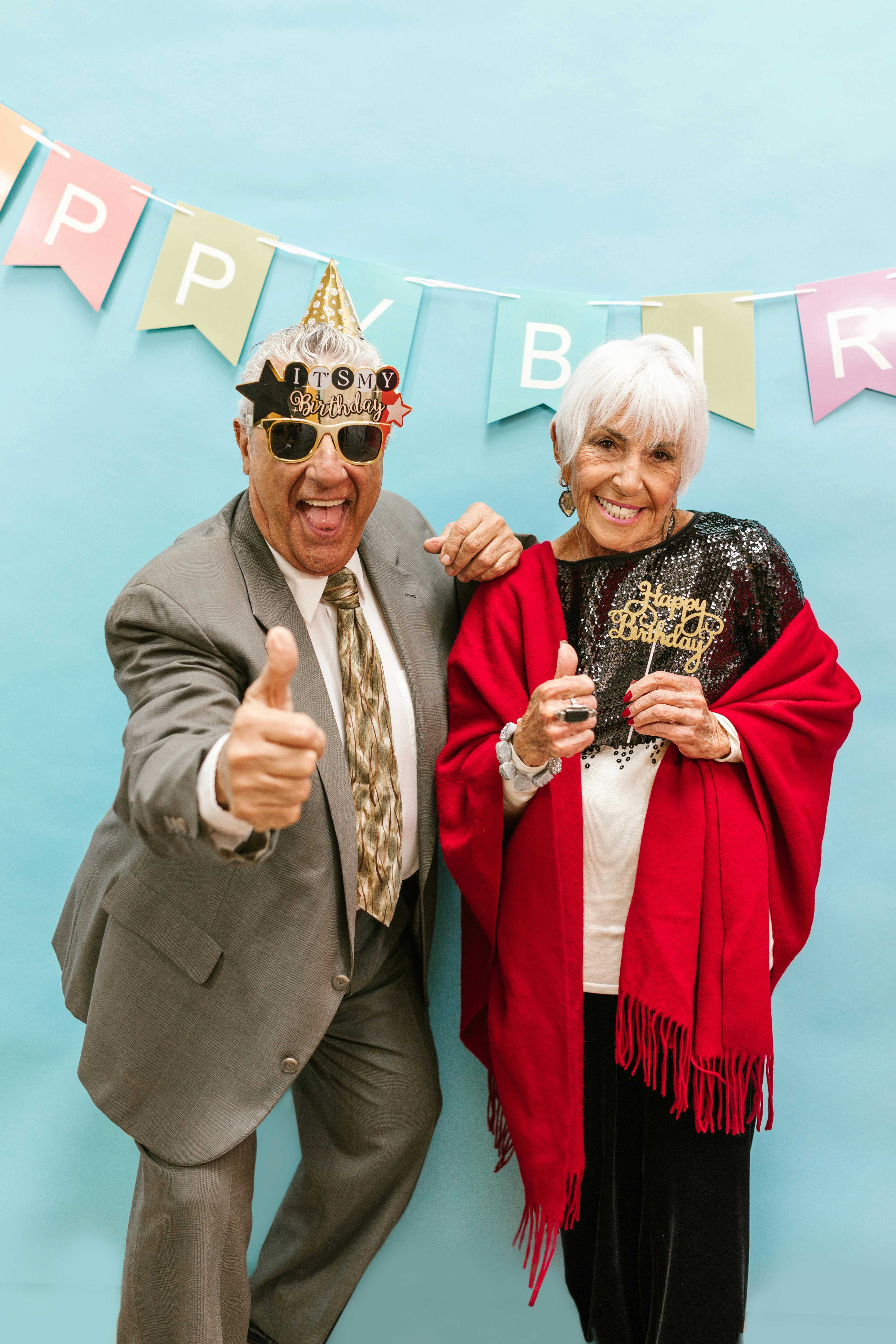 Smiling senior couple in festive attire celebrating a birthday with thumbs up and decorations.