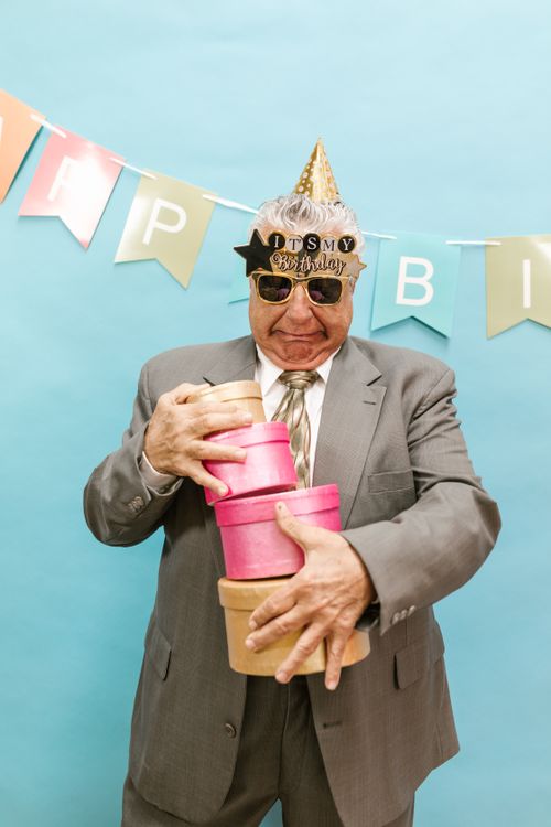 Free Elderly man in suit with party hat and sunglasses holds gift boxes against light blue background. Stock Photo