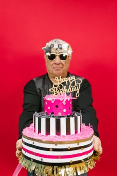 Senior man holding colorful tiered birthday cake against red background, wearing fun glasses.