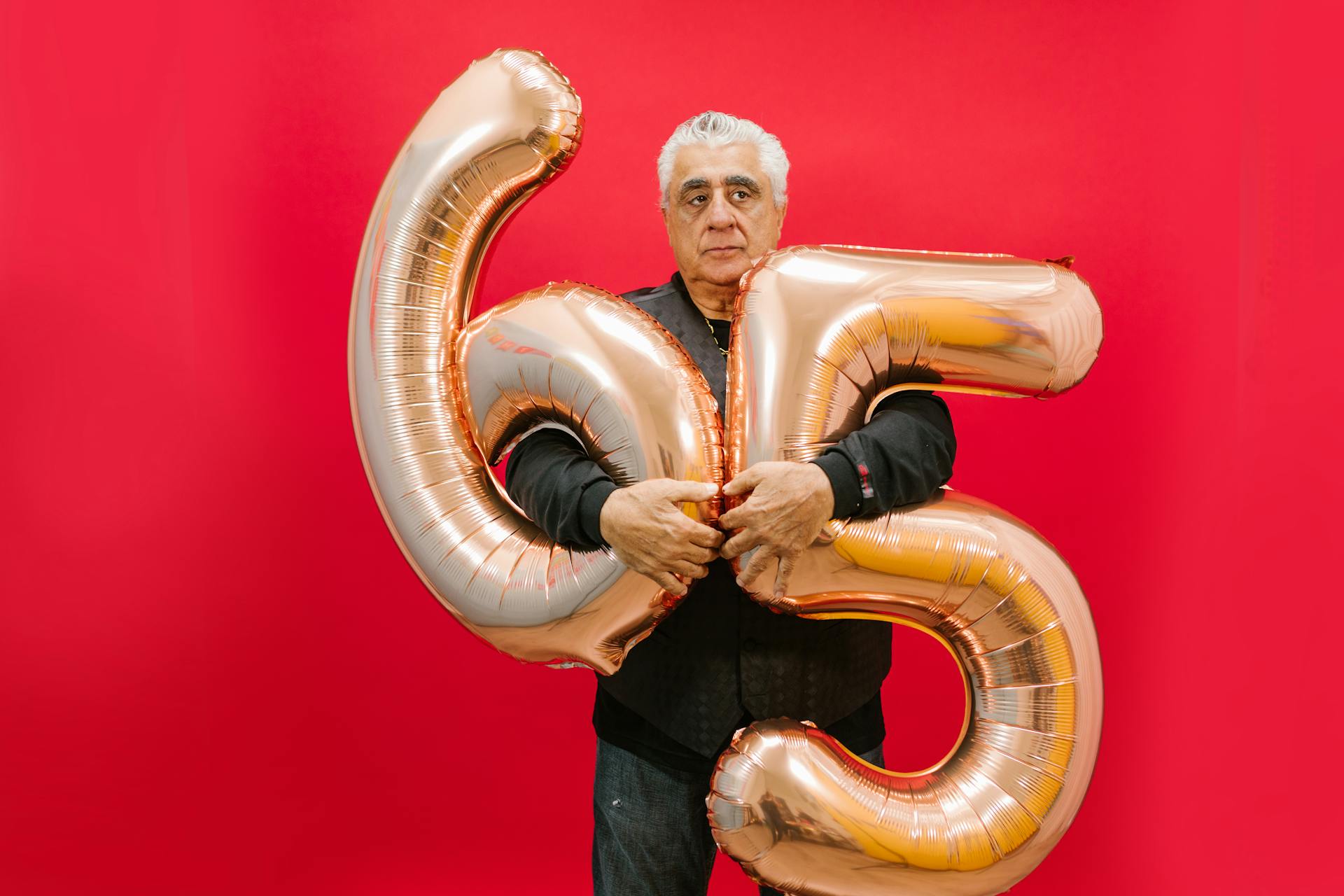 Senior man holding gold 65 number balloons against a red background.