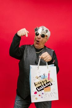 Cheerful senior man in sunglasses celebrating his birthday with a gift bag in a studio.
