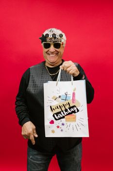 An elderly man celebrating his birthday holding a decorated gift bag against a vibrant red background.