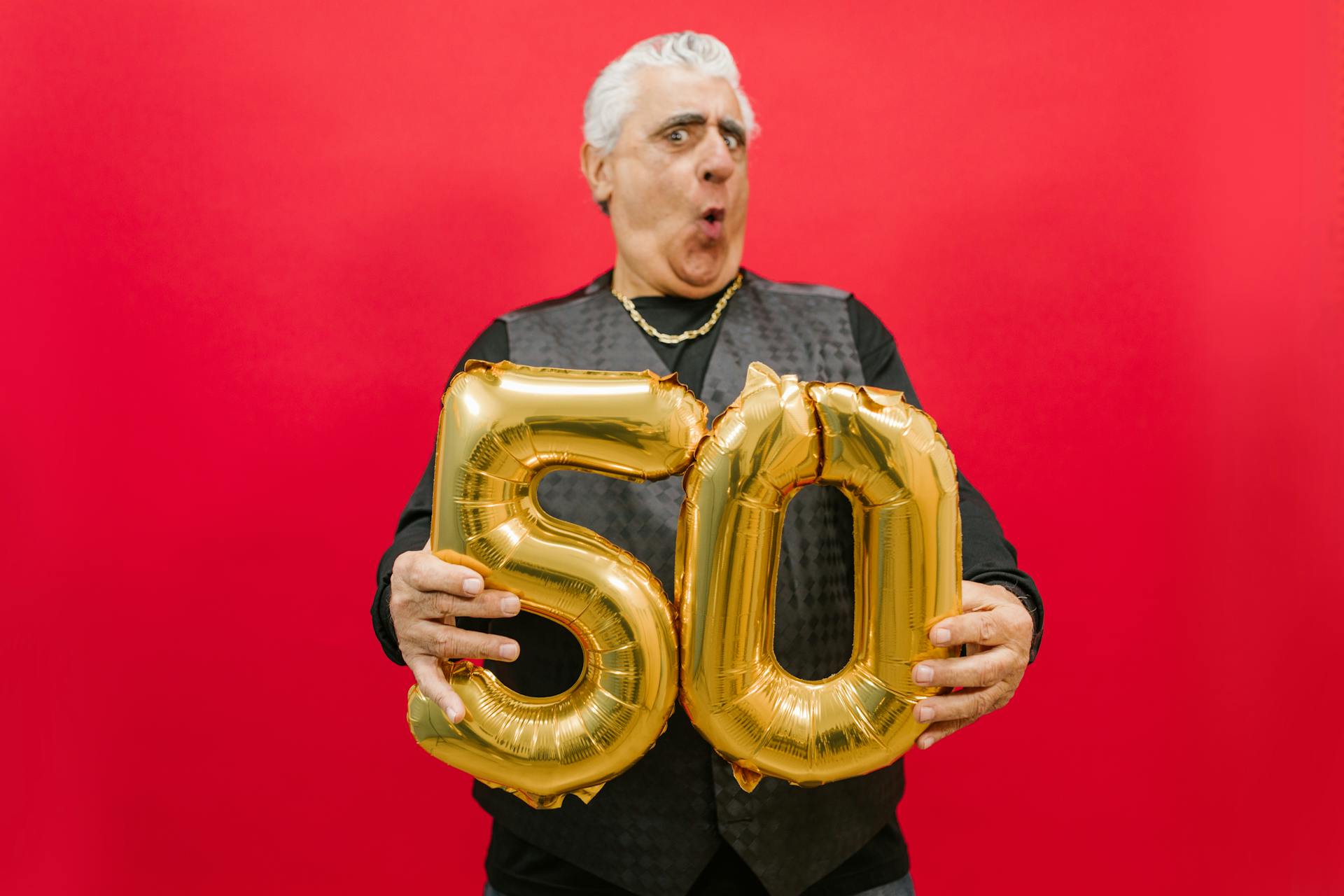 A joyful senior holding gold '50' balloons against a vibrant red backdrop.