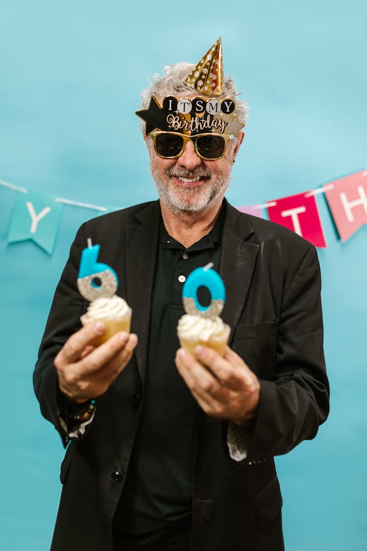 An Elderly Man Wearing Sunglasses And Party Hat While Holding A Cupcakes