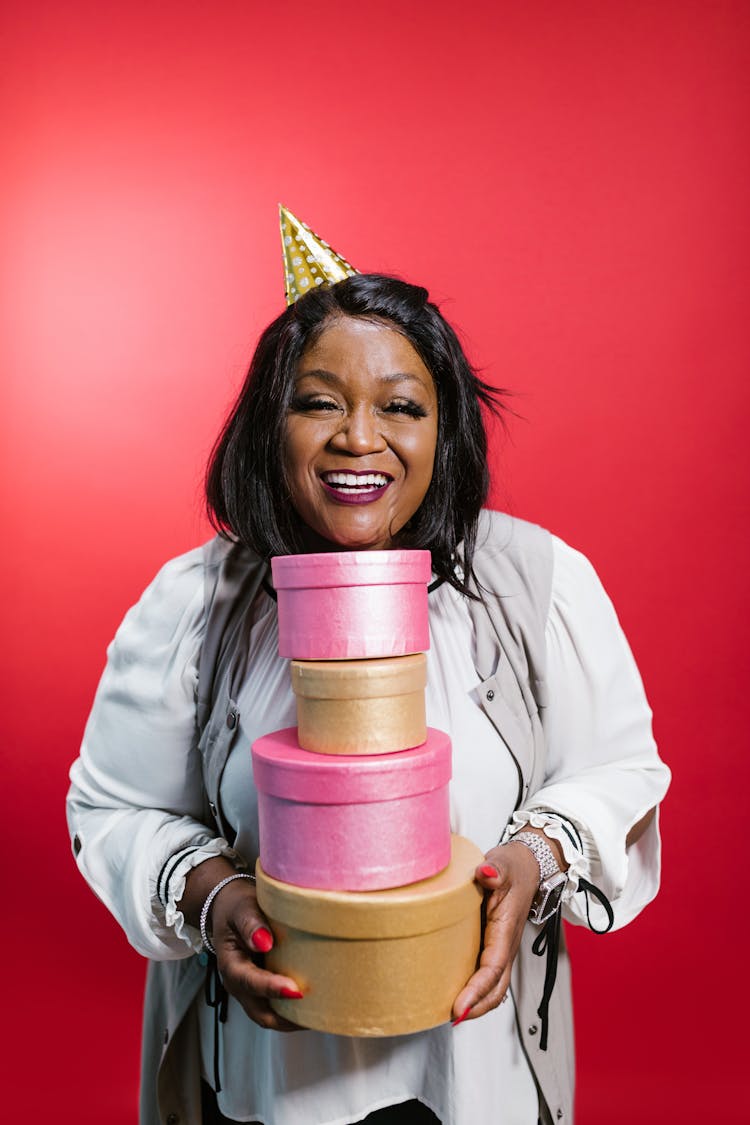 A Woman Wearing A White Top And Party Hat Holding A Pile Of Gifts