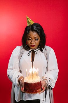 African American woman in a white top holding a birthday cake with lit candles against a red background.
