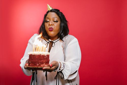 Free A black adult woman joyfully blowing out birthday candles on a red velvet cake against a red backdrop. Stock Photo
